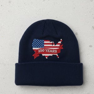 May include: A navy blue beanie featuring an embroidered design of the United States map in red, white, and blue, resembling the American flag. The design includes the text "250 YEARS" on a red banner.
