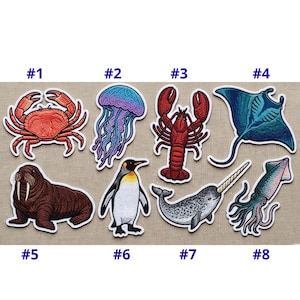 May include: Eight embroidered patches depicting marine life. The patches include a red crab, a jellyfish with purple and blue colours, a red lobster, a blue manta ray, a brown walrus, a penguin, a narwhal, and a squid.