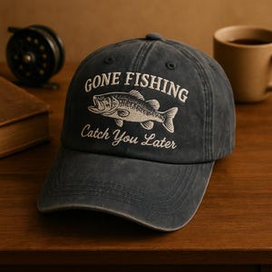 May include: A dark gray baseball cap with the text "GONE FISHING" above an embroidered fish graphic and the text "Catch You Later". The cap is made of a soft material and has a curved brim.
