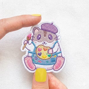 May include: Embroidered patch of a cartoon hamster artist. The hamster wears a purple beret and blue overalls, holding a paintbrush and a palette with heart-shaped paint. The patch has a white border. Ideal for clothing.