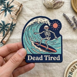 May include: Embroidered patch featuring a skeleton surfer riding a wave under a setting sun. The patch is navy blue with teal, orange, and white details. Text at the bottom reads "Dead Tired".