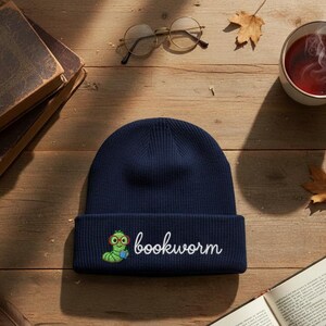 May include: A navy blue knit beanie with the word "bookworm" embroidered in white. The beanie features a cartoon bookworm with glasses and a book. The beanie is on a wooden surface with books and a cup of tea.