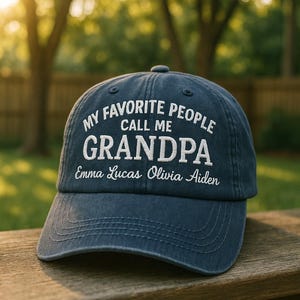 May include: A navy blue baseball cap with white embroidered text. The text reads "MY FAVORITE PEOPLE CALL ME GRANDPA" above the names Emma Lucas Olivia Aiden. The cap is sitting on a wooden surface.
