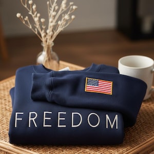 Peut inclure: Sweat-shirt bleu marine avec le mot « FREEDOM » brodé en blanc. La manche présente un écusson du drapeau américain. Le sweat-shirt est plié sur une surface tissée.