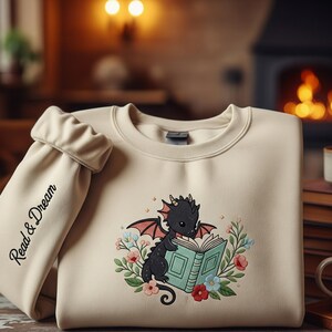 May include: Cream-colored sweatshirt featuring a black dragon reading a book, surrounded by flowers. The left sleeve has the words "Read & Dream" in black script. The design is a whimsical illustration.