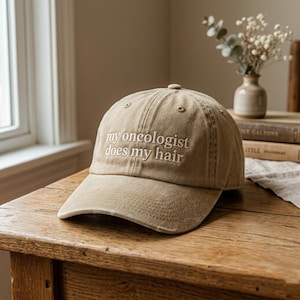 May include: A tan baseball cap with the embroidered text "my oncologist does my hair". The hat is made of a soft fabric and has a curved brim. The hat is sitting on a wooden surface.