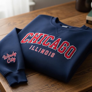 May include: A navy blue sweatshirt with the word "CHICAGO" in red and white lettering across the chest. The word "ILLINOIS" is below the city name. The left sleeve has the words "Windy City" embroidered in a cursive font.