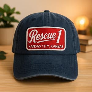 May include: A navy blue baseball cap with a red and white patch that reads "Rescue 1 Kansas City, Kansas". The cap is made of a soft, worn material and has a curved brim. The patch is rectangular with a white border.
