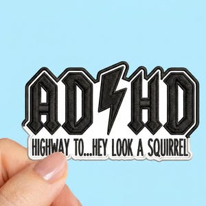 May include: Black and white embroidered patch with the text "ADHD" and a lightning bolt, styled like a rock band logo. Below, the text reads "HIGHWAY TO...HEY LOOK A SQUIRREL". The patch is held against a light blue background.
