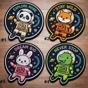 May include: Four embroidered patches with space-themed animal designs. Each patch features a different animal astronaut: a panda, fox, bunny, and dinosaur. The patches have text that reads "Explore More", "Stay Wild", "Dream Big", and "Never Stop".