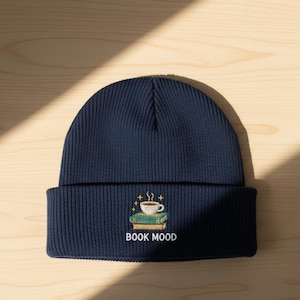 May include: Navy blue knit beanie with a folded brim. The front features an embroidered design of a coffee cup atop two books, with the words "BOOK MOOD" below. The beanie is made of a ribbed knit material.