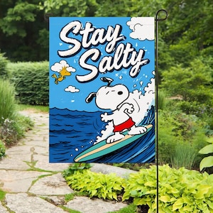 Może przedstawiać: Niebieska flaga ogrodowa z napisem "Stay Salty" w białym kolorze. Flaga przedstawia Snoopy'ego surfującego na fali, ubranego w czerwone kąpielówki. Żółty ptak znajduje się w lewym górnym rogu. Dekoracja ogrodu.