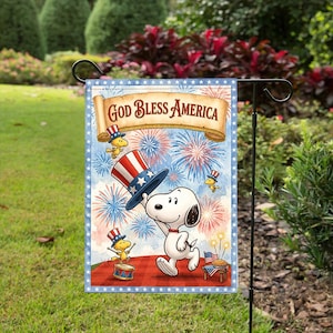 Może przedstawiać: Dekoracyjna flaga ogrodowa z Snoopy trzymającym patriotyczny kapelusz, z fajerwerkami i napisem "God Bless America". Flaga ma niebieską obwódkę z białymi gwiazdami i jest na czarnym metalowym stojaku.