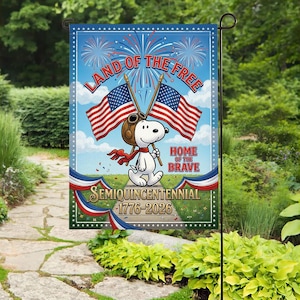 Może przedstawiać: Flaga ogrodowa ze Snoopy w czapce pilota trzymającym dwie amerykańskie flagi. Flaga zawiera tekst "Land of the Free" i "Home of the Brave" z fajerwerkami oraz tekst "Semiquincentennial 1776-2026". Flaga jest prezentowana w ogrodzie.