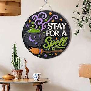 May include: A round black wooden wall sign featuring a gray cauldron with green bubbling liquid, a purple crescent moon, and a broomstick. The text reads STAY FOR A SPELL in white and green lettering, accented by small orange and purple stars.