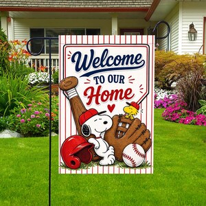 Könnte beinhalten: Gartenflagge mit Baseball-Motiv und dem Text "Welcome To Our Home". Die Flagge zeigt Snoopy, Woodstock, einen Baseballschläger, einen Handschuh, einen Helm und einen Baseball. Der Hintergrund hat rot-weiße Streifen.