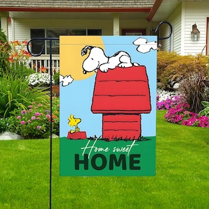 Może przedstawiać: Flaga ogrodowa przedstawiająca Snoopy'ego na czerwonej budzie i Woodstocka. Flaga ma niebieskie niebo, żółte słońce i napis "Home sweet HOME" w białym kolorze. Dekoracja ogrodu.