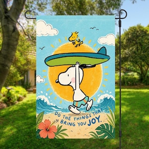 Może przedstawiać: Dekoracyjna flaga ogrodowa z ilustracją Snoopy'ego niosącego deskę surfingową, z Woodstockiem na górze, na tle żółtego słońca i niebieskich fal. Flaga zawiera napis "Do the things that bring you joy."