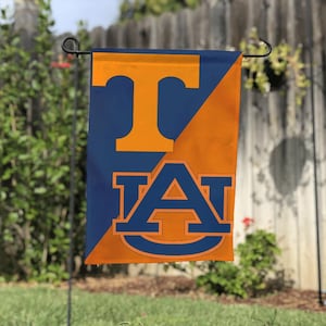 Puede incluir: Una bandera de jardín azul y naranja con la letra "T" en naranja en la parte superior y las letras "A" y "T" en azul y naranja en la parte inferior. La bandera está colgada al aire libre en un poste de metal.