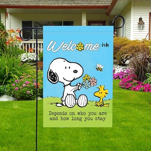 Może przedstawiać: Niebieska flaga ogrodowa z Snoopy'm trzymającym kwiaty, Woodstockiem i pszczołą. Flaga zawiera napis "Welcome-ish" i frazę "Depends on who you are and how long you stay."