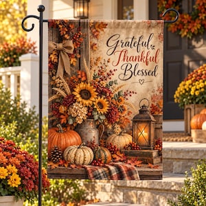 May include: A rectangular garden flag featuring the text Grateful Thankful Blessed, a rustic metal pitcher filled with sunflowers, wheat, and autumn leaves, a lit lantern, pumpkins, pinecones, and a plaid cloth on a wooden surface.