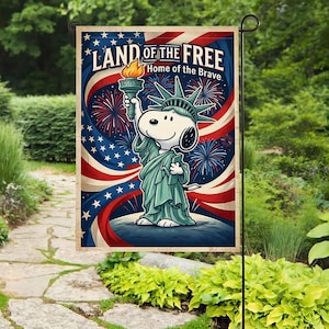 Może przedstawiać: Flaga ogrodowa przedstawiająca Snoopy'ego jako Statuę Wolności, trzymającego pochodnię. Flaga ma patriotyczny wzór z napisem "LAND OF THE FREE, Home of the Brave" oraz czerwoną, białą i niebieską kolorystykę z fajerwerkami i gwiazdami.