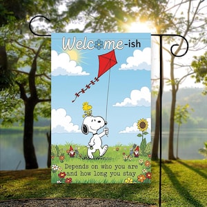Może przedstawiać: Flaga ogrodowa z Snoopy'm z Peanuts, trzymającym czerwoną latawiec z Woodstockiem na głowie. Flaga ma napis "Welcome-ish" i "Depends on who you are and how long you stay" na tle błękitnego nieba.