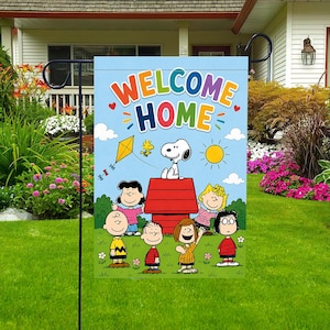 Może przedstawiać: Niebieska flaga ogrodowa z napisem "WELCOME HOME" w kolorowych literach. Flaga przedstawia Snoopy'ego, Woodstocka i inne postacie z Peanuts w słonecznej scenerii na świeżym powietrzu z latawcem i czerwonym budą.