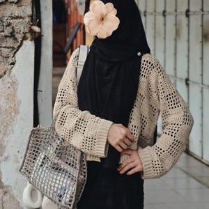 Pu&ograve; includere: Un cardigan beige all'uncinetto a trama aperta con maniche lunghe. Il cardigan &egrave; abbinato a un hijab e una gonna neri. Una borsa a rete grigia contiene oggetti e cuffie bianche sono appese alla borsa.