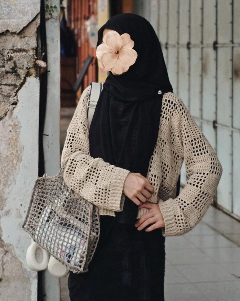 Pu&ograve; includere: Un cardigan beige all'uncinetto a trama aperta con maniche lunghe. Il cardigan &egrave; abbinato a un hijab e una gonna neri. Una borsa a rete grigia contiene oggetti e cuffie bianche sono appese alla borsa.