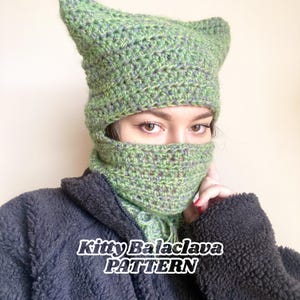 Può includere: Una balaclava a forma di gattino in crochet verde e blu con orecchie da gatto. La balaclava copre la testa e il viso, solo gli occhi sono visibili. Il testo "Kitty Balaclava PATTERN" è nella parte inferiore dell'immagine.