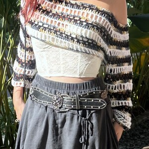 May include: A white lace corset top with a black and white striped off-the-shoulder crochet sweater. The ensemble is finished with a dark grey skirt and a black studded belt with a decorative buckle.
