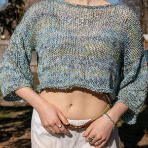 May include: A cropped, open-weave sweater in shades of blue, green, and brown. The sweater has a wide neckline and three-quarter sleeves. The model is wearing a white skirt and a brown belt. The sweater is a handmade item.