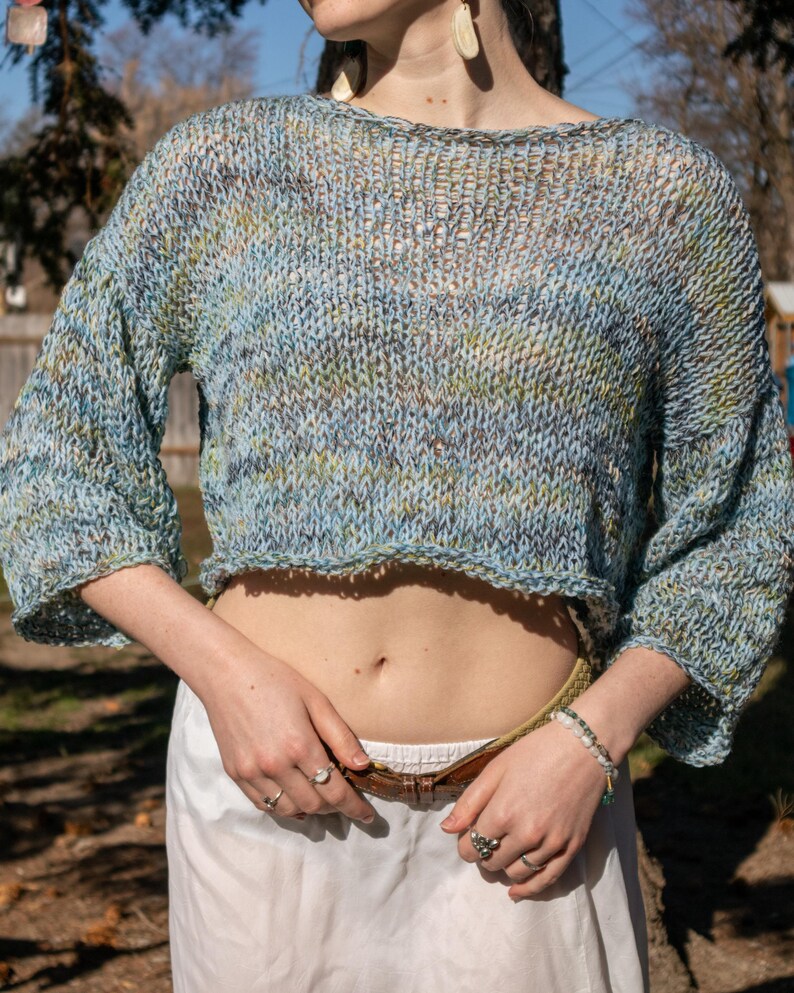 May include: A cropped, open-weave sweater in shades of blue, green, and brown. The sweater has a wide neckline and three-quarter sleeves. The model is wearing a white skirt and a brown belt. The sweater is a handmade item.
