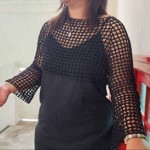 May include: A black crochet top with long sleeves. The top is worn over a black dress. The crochet design features a grid-like pattern. The sleeves are flared at the wrist. The top is a stylish addition to any wardrobe.