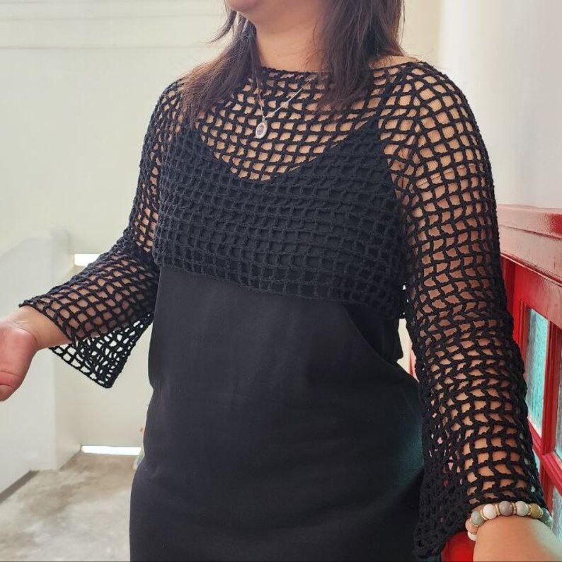 May include: A black crochet top with long sleeves. The top is worn over a black dress. The crochet design features a grid-like pattern. The sleeves are flared at the wrist. The top is a stylish addition to any wardrobe.