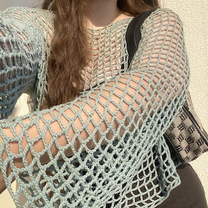 May include: A pale blue crochet top with long sleeves. The open weave design creates a net-like pattern. The top is worn with a black bag with a brown and beige pattern.