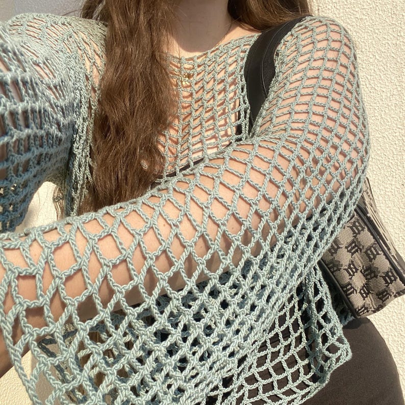 May include: A pale blue crochet top with long sleeves. The open weave design creates a net-like pattern. The top is worn with a black bag with a brown and beige pattern.