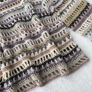 May include: A hand-crocheted sweater in a neutral colour palette. The open-weave design features stripes of beige, grey, and purple. The sleeves are long and the sweater has a relaxed fit. The yarn appears to be soft and textured.