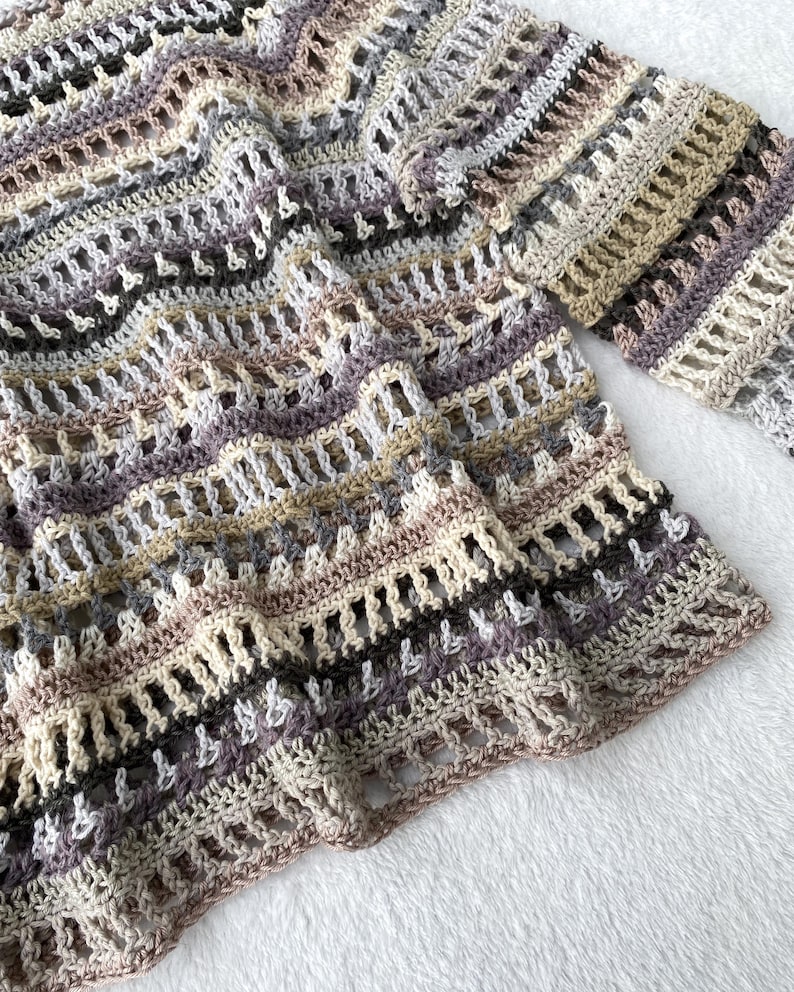 May include: A hand-crocheted sweater in a neutral colour palette. The open-weave design features stripes of beige, grey, and purple. The sleeves are long and the sweater has a relaxed fit. The yarn appears to be soft and textured.