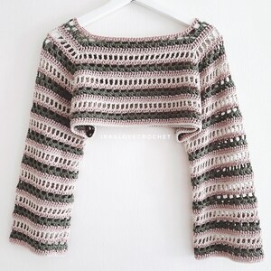May include: Crochet crop top with a striped design. The top has long sleeves and a boat neck. The colour scheme includes shades of grey, pink, and white. The garment is displayed on a white hanger against a white background.