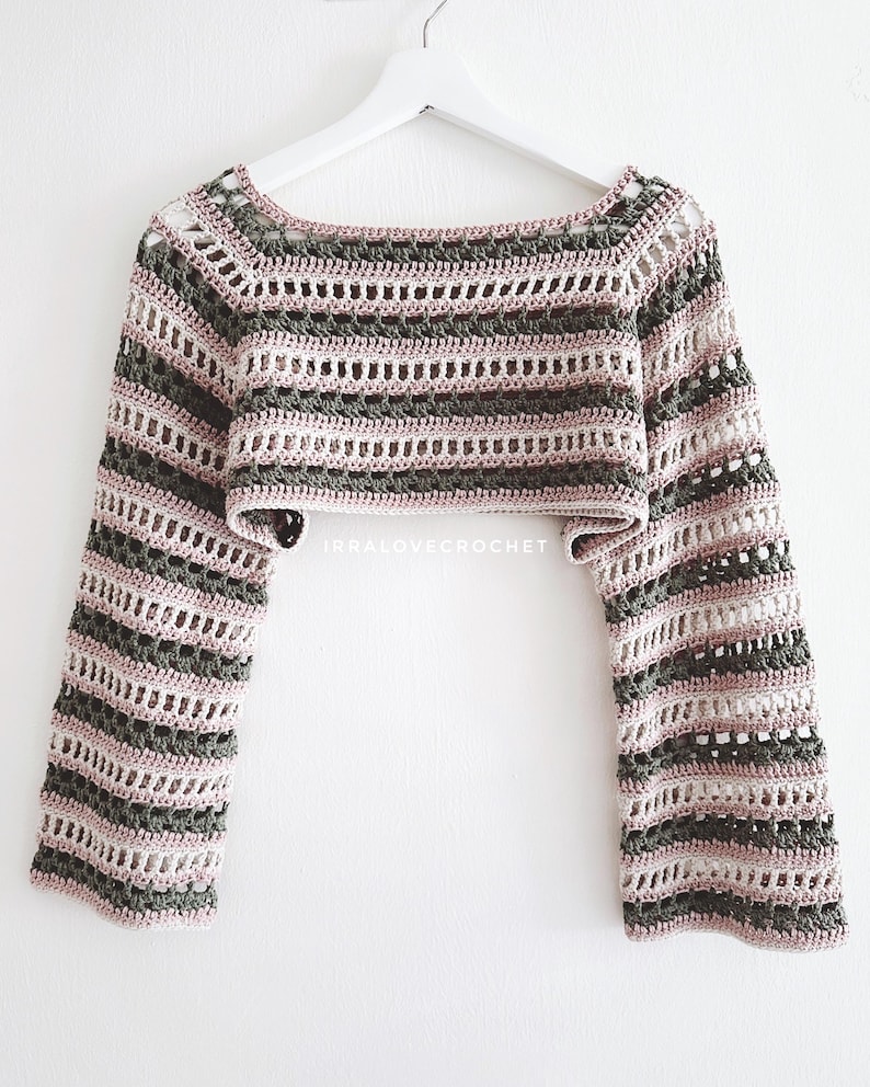 May include: Crochet crop top with a striped design. The top has long sleeves and a boat neck. The colour scheme includes shades of grey, pink, and white. The garment is displayed on a white hanger against a white background.