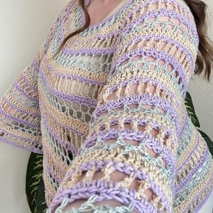 May include: A pastel-coloured crochet top with a scoop neck and long sleeves. The top features a striped pattern in shades of lavender, cream, and light blue. The open weave design adds a delicate and airy feel to the garment.