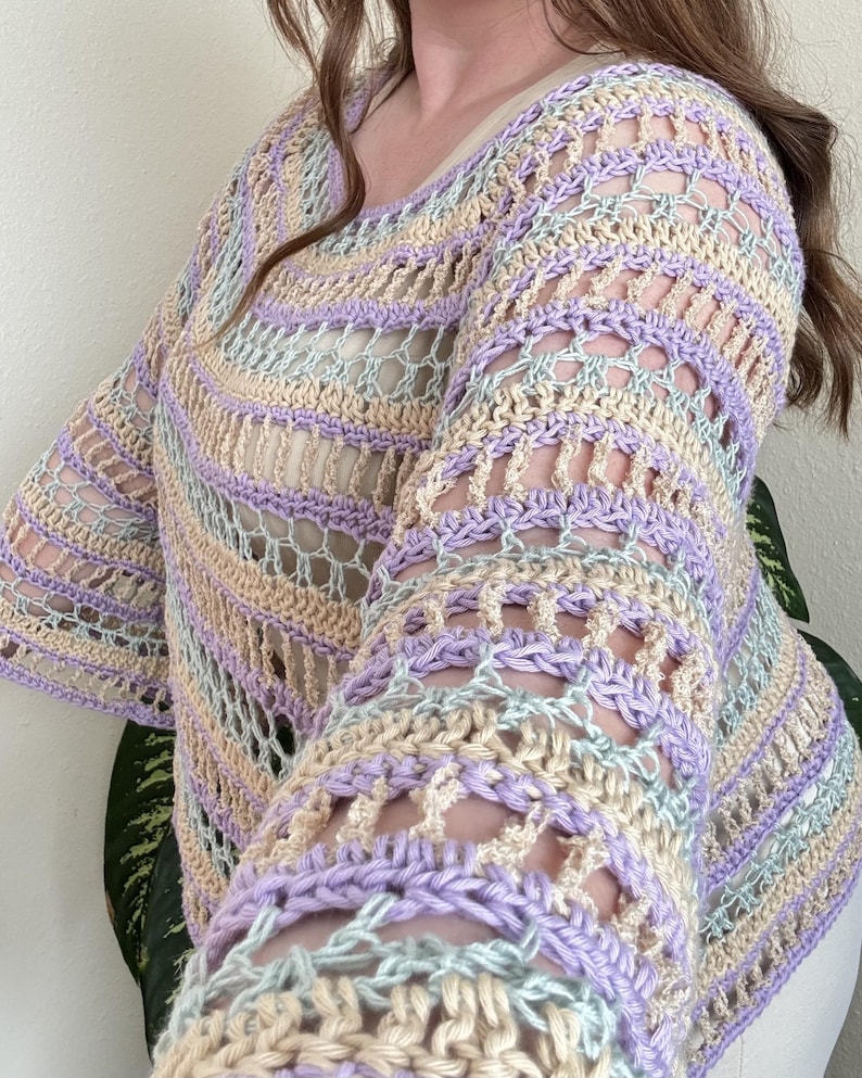 May include: A pastel-coloured crochet top with a scoop neck and long sleeves. The top features a striped pattern in shades of lavender, cream, and light blue. The open weave design adds a delicate and airy feel to the garment.