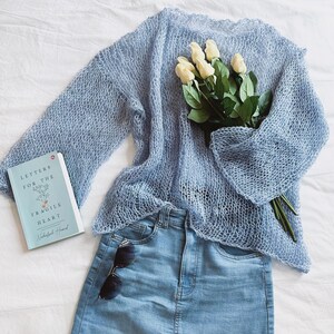 May include: A light blue, open-weave sweater with wide sleeves is paired with light wash denim jeans. A bouquet of white roses and a book titled "Letters for the Fragile Heart" are also present. Sunglasses rest on the jeans.