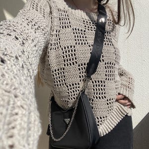 Pu&ograve; includere: Un maglione beige all'uncinetto a trama aperta con maniche lunghe. Una borsa a tracolla nera con una tracolla a catena argentata &egrave; appesa alla spalla. La borsa ha un piccolo logo argentato. La persona indossa pantaloni neri.