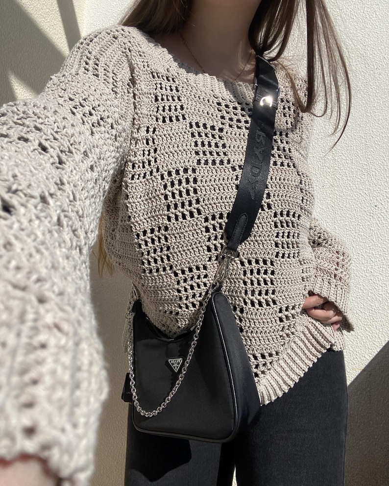 Pu&ograve; includere: Un maglione beige all'uncinetto a trama aperta con maniche lunghe. Una borsa a tracolla nera con una tracolla a catena argentata &egrave; appesa alla spalla. La borsa ha un piccolo logo argentato. La persona indossa pantaloni neri.