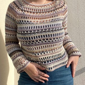 May include: A long-sleeved, open-weave crochet top in a neutral colour palette of beige, lavender, and grey. The top features a round neckline and a slightly flared sleeve. The crochet design creates a textured, airy look.