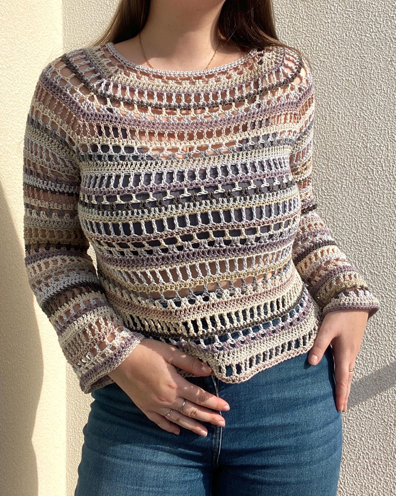 May include: A long-sleeved, open-weave crochet top in a neutral colour palette of beige, lavender, and grey. The top features a round neckline and a slightly flared sleeve. The crochet design creates a textured, airy look.