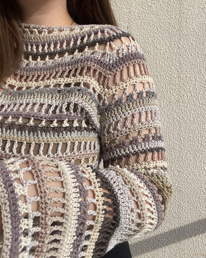 May include: A close-up of a crochet top in neutral tones. The open-weave design features stripes of beige, grey, and brown. The long sleeves and high neckline are visible. The top appears to be handmade.